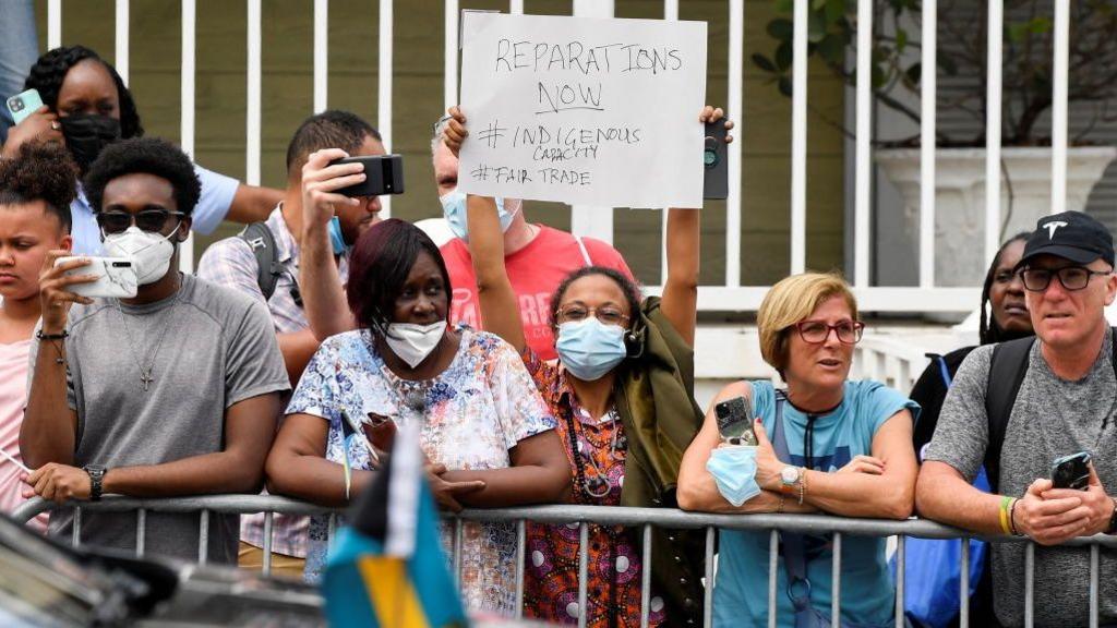 Image caption,A woman holds a sign in protest as she waits for the arrival of William and Catherine on their tour