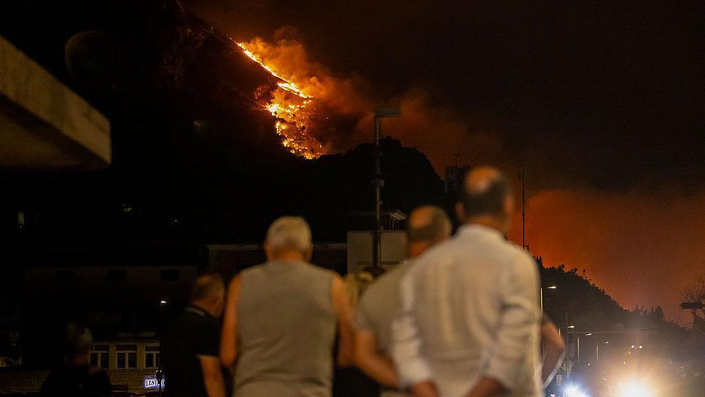 Image caption,Bystanders watch as a wooden area is engulfed in flames during a raging wildfire near the town of Omis