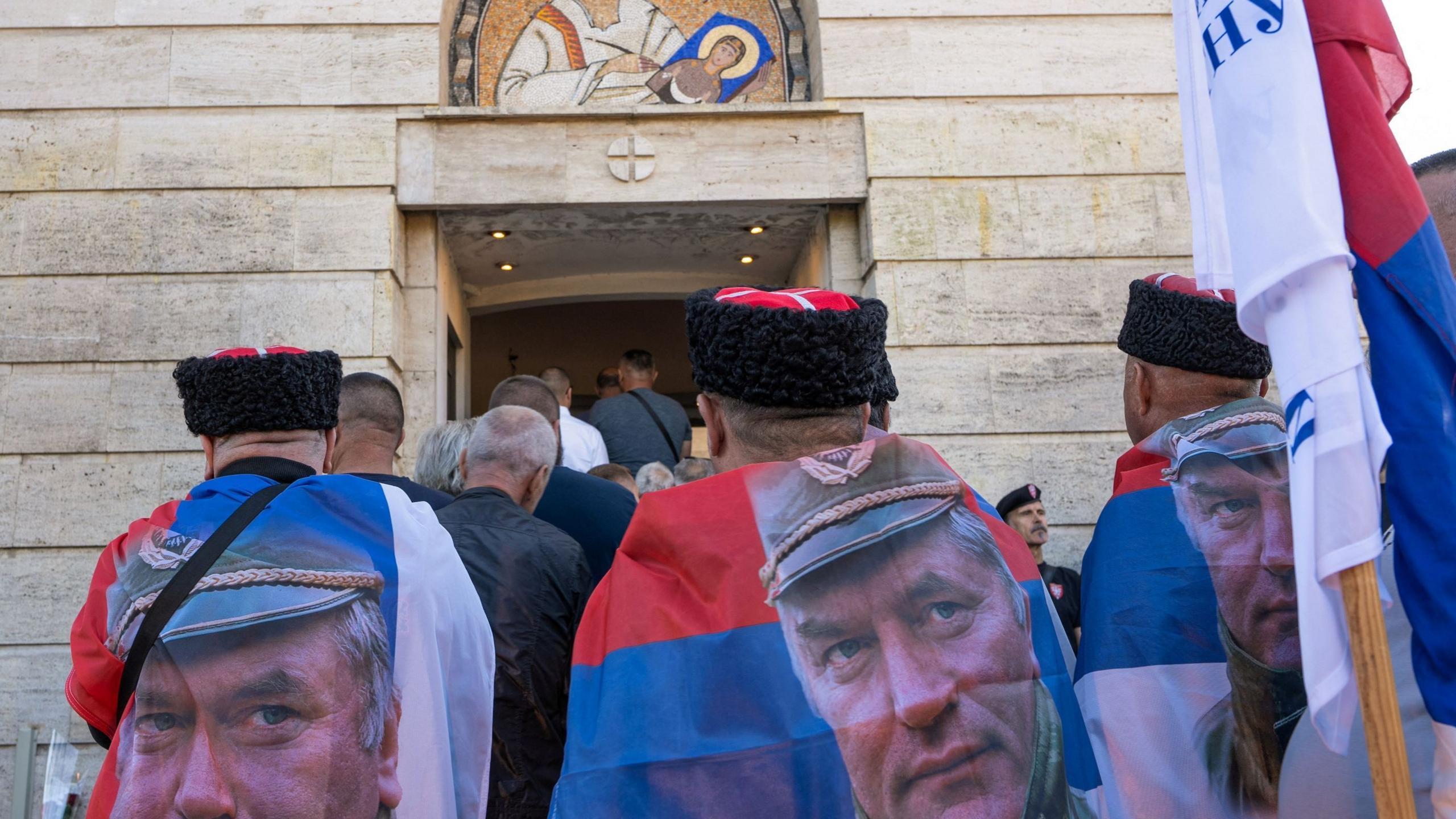 Image caption,Men wearing flags as capes, with the picture of Mladić, stood among the crowd in Belgrade