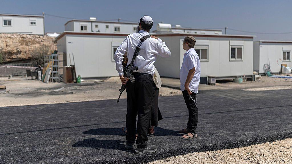 Image caption,Three Israeli settlers - including an armed one - stand outside houses after the reopening of a settlement at Ganim in the Israeli-occupied West Bank in August